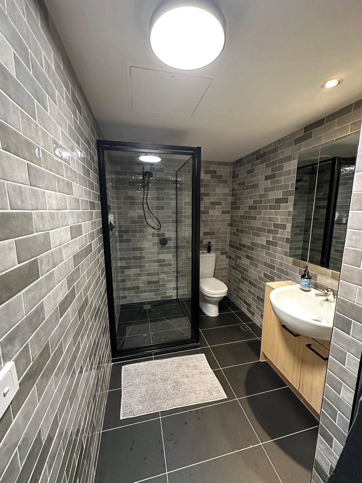 Bathroom with grey brick-format wall tiles, dark floor and black shower screen